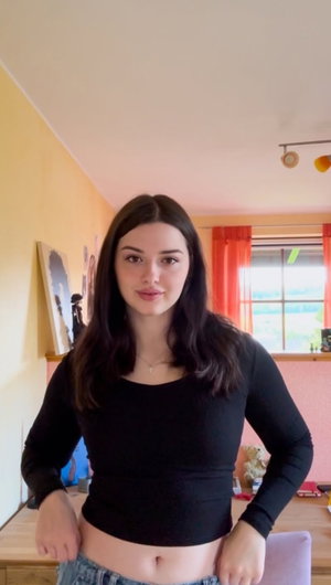 Woman with dark hair in black long-sleeve crop top and jeans, standing indoors.