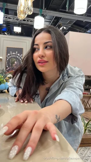 Woman with dark hair and tattoos wears denim shirt, leaning on counter in indoor setting.