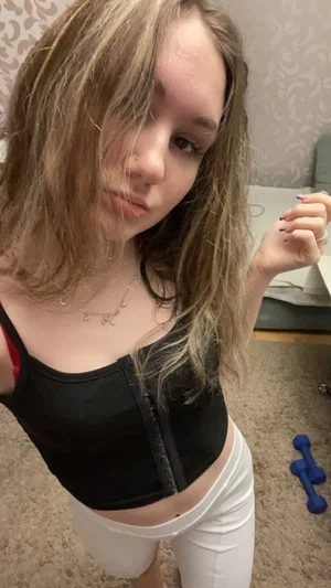 Woman with blonde hair in black crop top and white pants, posing indoors.