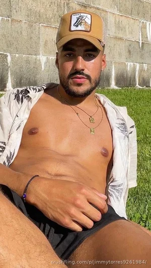 Man with beard and cap, shirt open, sitting outdoors on grass near concrete wall.