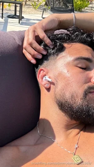 Shirtless man with curly hair and beard resting outdoors, wearing earbuds and necklace.