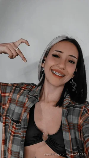 Woman with two-toned hair, wearing black top and plaid shirt, smiling in indoor setting.