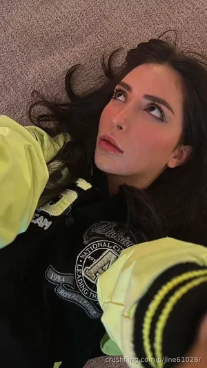 Woman with dark hair lying on carpet, wearing black jacket with yellow sleeves and patches.