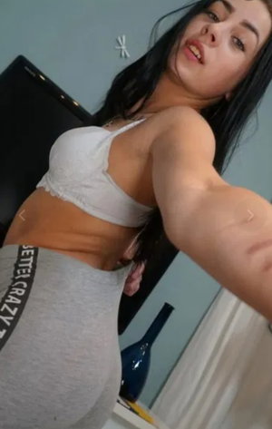 Woman with long dark hair in white bra and grey pants taking selfie indoors.