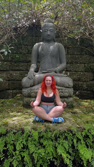 Woman with red hair sits cross-legged in front of stone Buddha statue in a lush green garden.