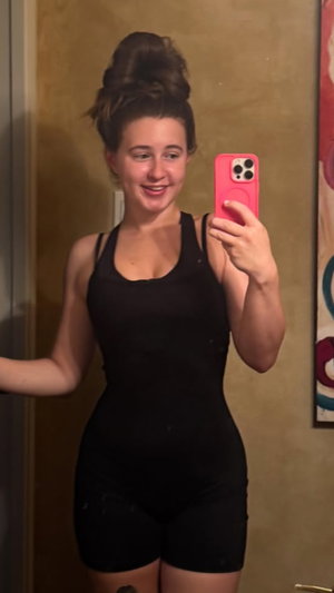 Woman in black one-piece outfit taking mirror selfie indoors.