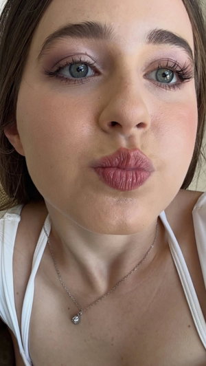 Woman with blue eyes and makeup, wearing white top and necklace, making kiss face.