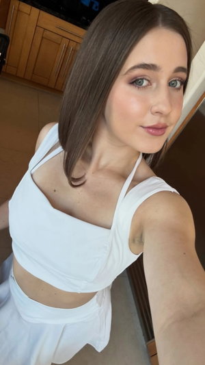 Woman with brown hair in white crop top and skirt, taking selfie indoors.
