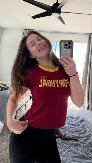 Woman in red Portugal t-shirt and black pants takes mirror selfie in bedroom.