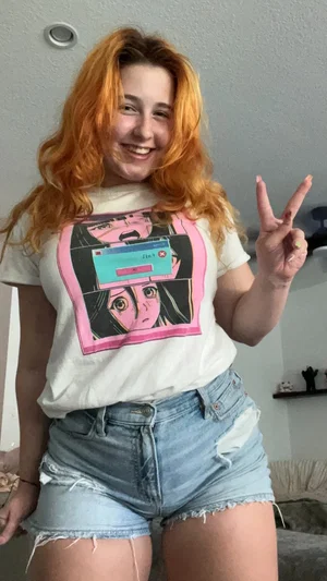 Woman with orange hair in white graphic tee and ripped denim shorts posing indoors.