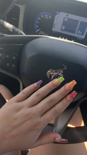 Hand with colorful nails resting on car steering wheel, interior visible.
