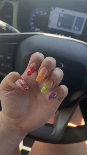 Hand with colorful fruit-themed nail art inside a car, showing steering wheel and dashboard.