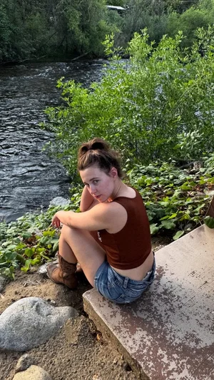 Woman in brown tank top and denim shorts sits by river, outdoor setting with greenery.