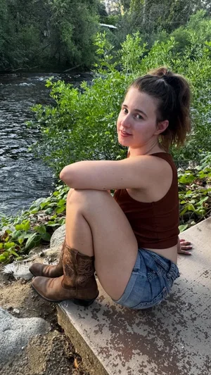 Woman in brown tank top and denim shorts sits by river, wearing cowboy boots.
