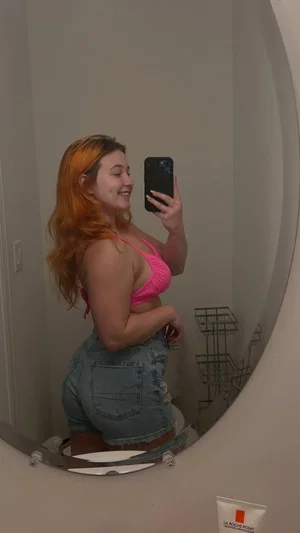 Woman with orange hair in pink top and denim shorts takes mirror selfie in bathroom.