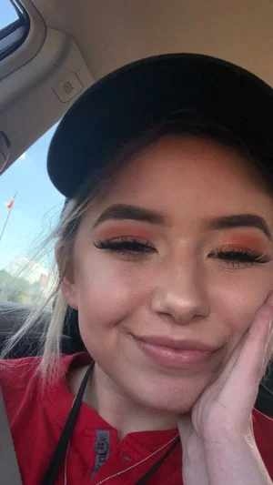 Woman with makeup and black cap, wearing red shirt, smiling in car.