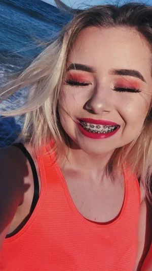 Woman with blonde hair and red lipstick wearing orange tank top, smiling outdoors near water.