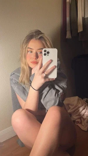 Woman with blonde hair in gray shirt taking mirror selfie indoors, legs bare.