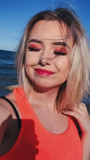 Woman with blonde hair and red makeup wearing orange tank top at beach.