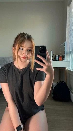 Woman in black t-shirt taking mirror selfie in bedroom, bare legs visible.