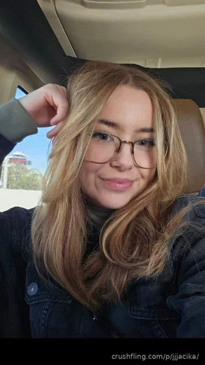 Woman with blonde hair and glasses, wearing a dark jacket, taking a selfie in a car.
