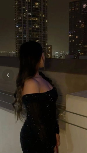 Woman in black sequined off-shoulder dress on rooftop at night, city lights behind.