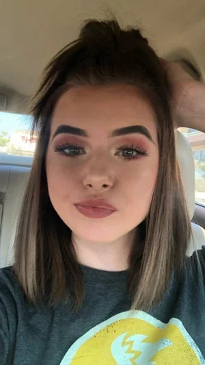 Woman with brown hair and makeup wearing a gray t-shirt in a car.