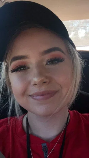 Woman with blonde hair and makeup wearing red shirt and black cap, smiling in car.
