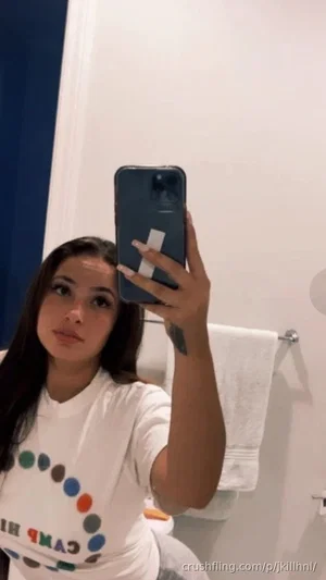 Woman with long dark hair in white t-shirt taking mirror selfie in bathroom.