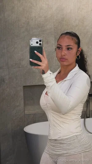 Woman in white long-sleeve top and light pants taking mirror selfie in bathroom.