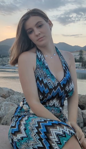 Woman in patterned halter dress sitting by waterfront at dusk, mountains in background.