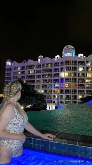 Woman in patterned swimsuit by pool at night, facing hotel building.