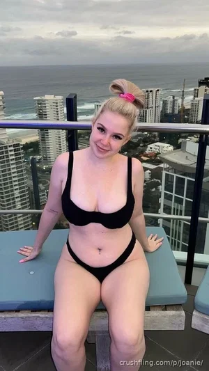 Woman in black bikini posing on rooftop with ocean and city view.
