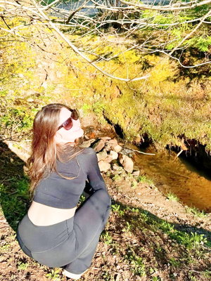 Woman in black crop top and leggings crouching by a stream in a wooded area.