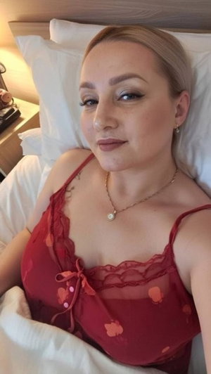 Woman with blonde hair in red lace lingerie lying in bed, wearing necklace.