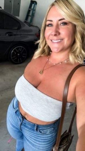 Blonde woman in gray crop top and jeans taking selfie in garage.