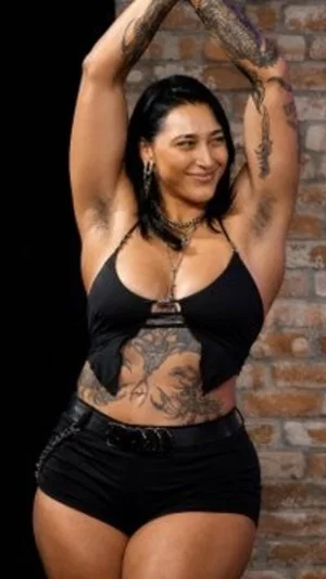 Woman with tattoos in black top and shorts posing in front of brick wall.