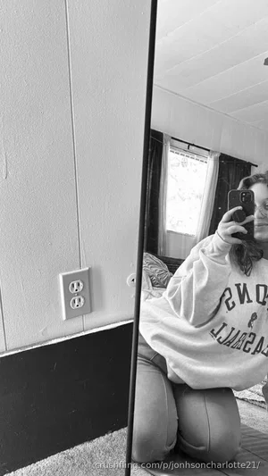 Woman in oversized sweatshirt and pants kneeling on floor, taking mirror selfie indoors.