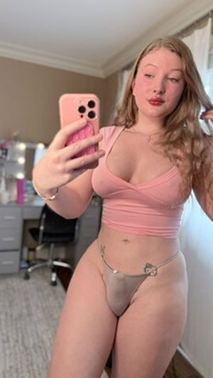 Woman in pink crop top and sheer thong takes mirror selfie in bedroom.
