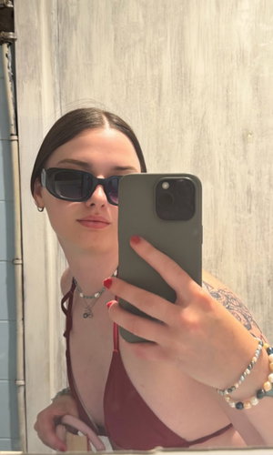Woman in maroon top with tattoos takes mirror selfie, wearing sunglasses and necklace.