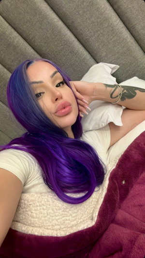 Curvy woman with purple hair in white top on bed.