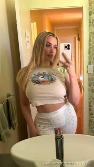 Blonde woman in crop top and patterned pants takes mirror selfie in bathroom.