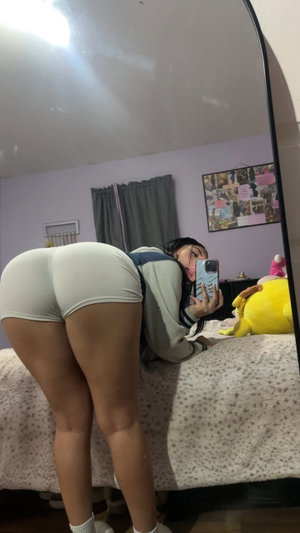 Woman in white shorts and long-sleeve top taking mirror selfie in bedroom.