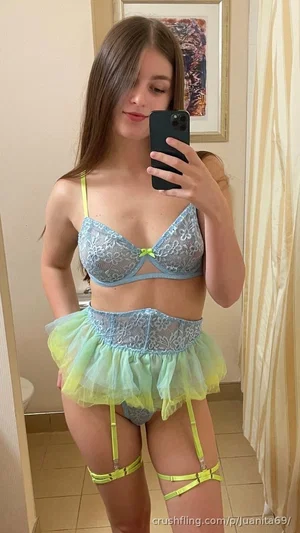 Woman in light blue lace bra and colorful garter belt, taking mirror selfie in indoor setting.