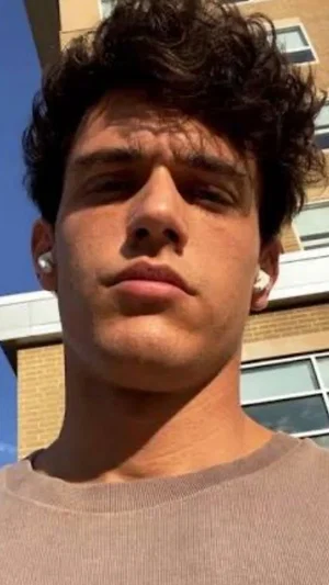 Man with curly dark hair wearing a brown shirt outdoors in front of a building.