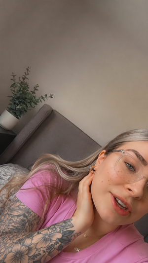 Blonde woman with glasses in pink shirt taking selfie indoors.