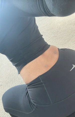 Woman in black workout outfit with visible navel piercings, kneeling on carpet.