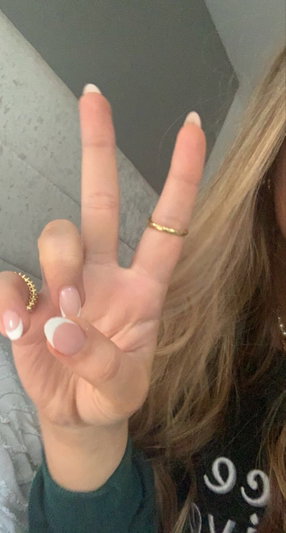Woman with blonde hair and manicured nails making peace sign, wearing dark green top indoors.
