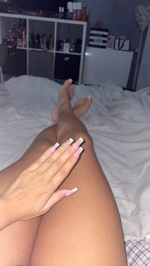 Woman with long nails sitting on bed, legs crossed, hand on thigh, indoor setting.