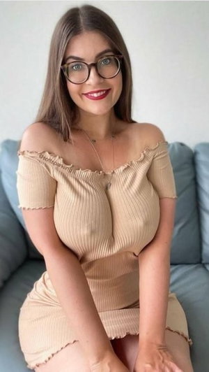 Woman with glasses in beige off-shoulder dress sitting on blue couch indoors.